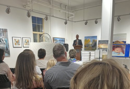 Joseph Gibbons stands before a seated crowd in an art gallery presenting his new book.