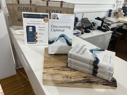White hard-copy books sit on a wooden slab with marketing signs in a well-lit room. 