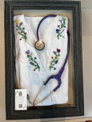 in a black picture frame, a stethoscope is wrapped around a white shirt, with colourful flowers embroidered onto it. The white button on the shirt has 'cheroke' engraved on it. 