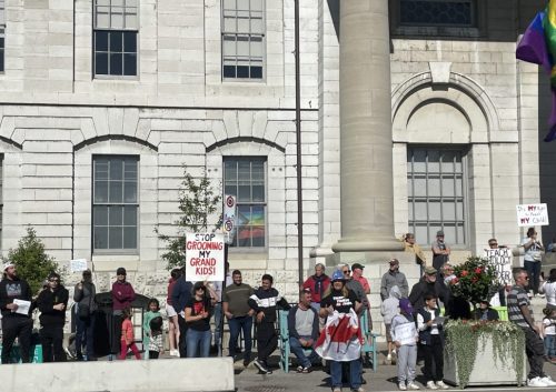 1 Million March protestors in front of city hall. A woman in the front is holding a canadian flag, another protestor behind her holds a sign that reads 'stop grooming our grandkids'. an lgbt+ flag is in the window inside City Hall.