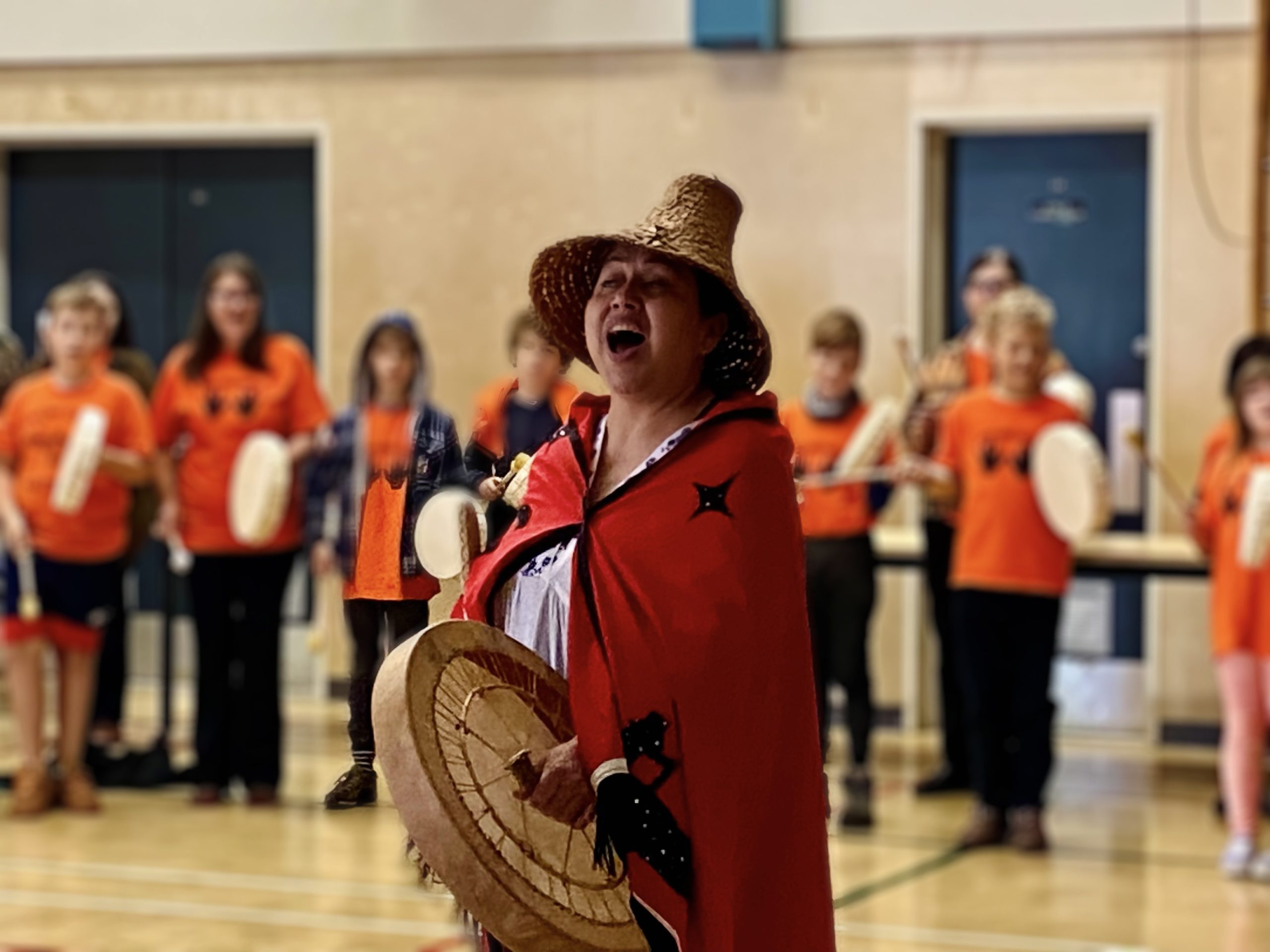 A women wrapped in orange, sings and plays a drum, leading a group of children behind her.
