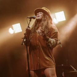  Shawnee Kish performing live on a stage. She stands at a microphone wearing a hat and there are floodlights behind her.