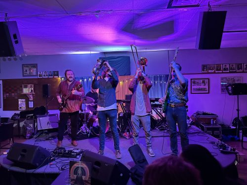 Typically a 9 piece ensemble, Six members of the band 'Apollo Suns' play their instruments on a Legion stage in Prince George BC. They point their instruments toward the ceiling in choreographed unison. A blue and purple light washes over them.