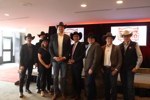 Dignitaries from Explore Edmonton, the City of Edmonton, Edmonton Destination Marketing Hotels, the Government of Alberta, the Canadian Professional Rodeo Association, stand in line front of the podium at the Edmonton Convention Centre. Photo was taken inside, with curtains and red lights behind the subjects.