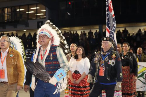 Lots of Indigenous peoples in regalia walking during the grand entry of the National Gathering of Elders. Photo was taken in a dark space.