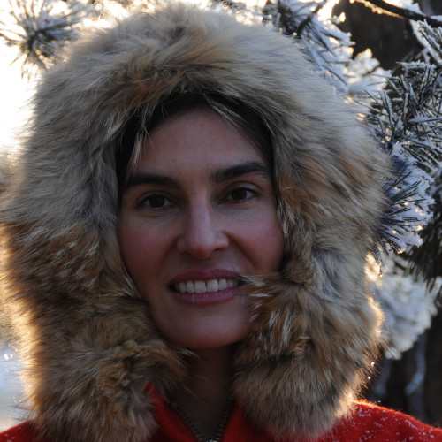 Michelle Connolly's head is framed in a outodoor winter image. She wers a fur lined hood pulled around her head, features red from cold. Red shoulders of jacket are visible. Snow covered pine branches are visibl in background.