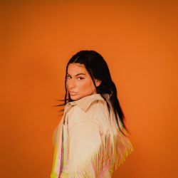  Shawnee Kish stands against an orange background in a professional cover photo. She has dark hair and wears a white jacket.