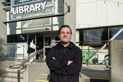 A man stands in front of a library with his arms crossed.