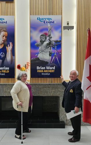 A woman and a man stand alongside a photo of an Olympic Para Archery athlete