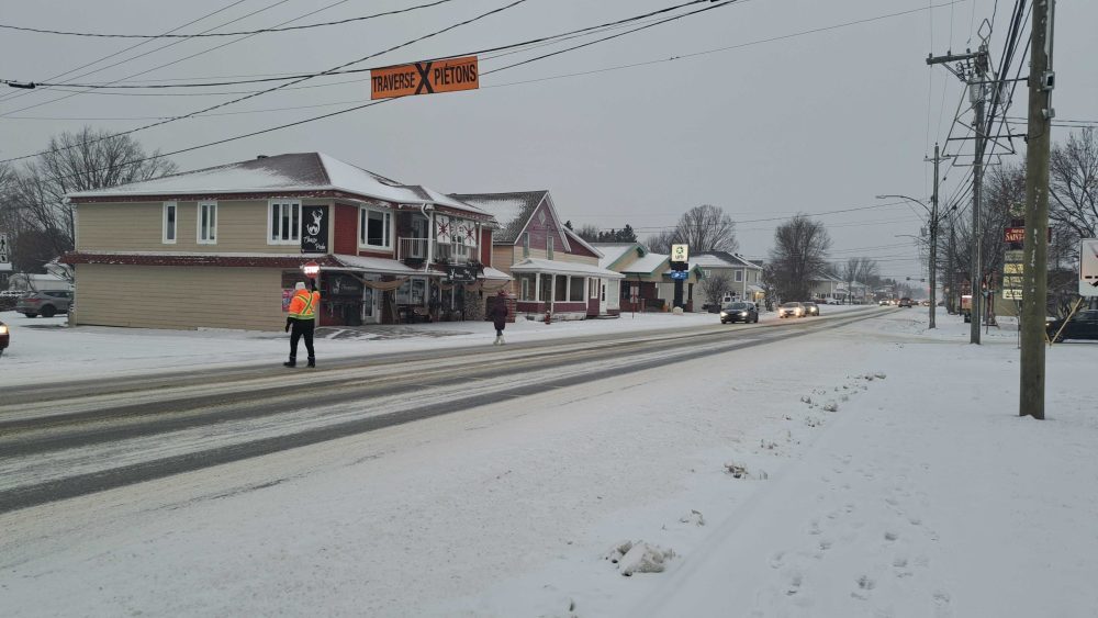 Une brigadière tenant une pancarte d'arrêt aidant une écolière à traverser une rue enneigée. Des véhicules sont arrêtés pour laisser passer l'écolière, lors d'une journée de novembre nuageuse et enneigée