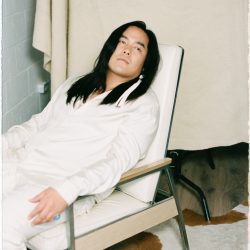 man siting in white chair, in all white outfit, with long black hair