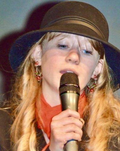Une jeune fille portant un chapeau brun et chantant dans un micro.