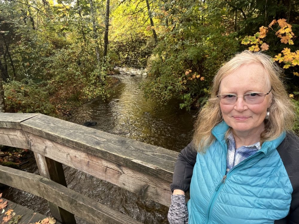 a blonde haired , smiling woman with glasses and a turquoise vest leaning on the rail of a bridge that looks to a long golden-green, treed Morrison Creek.