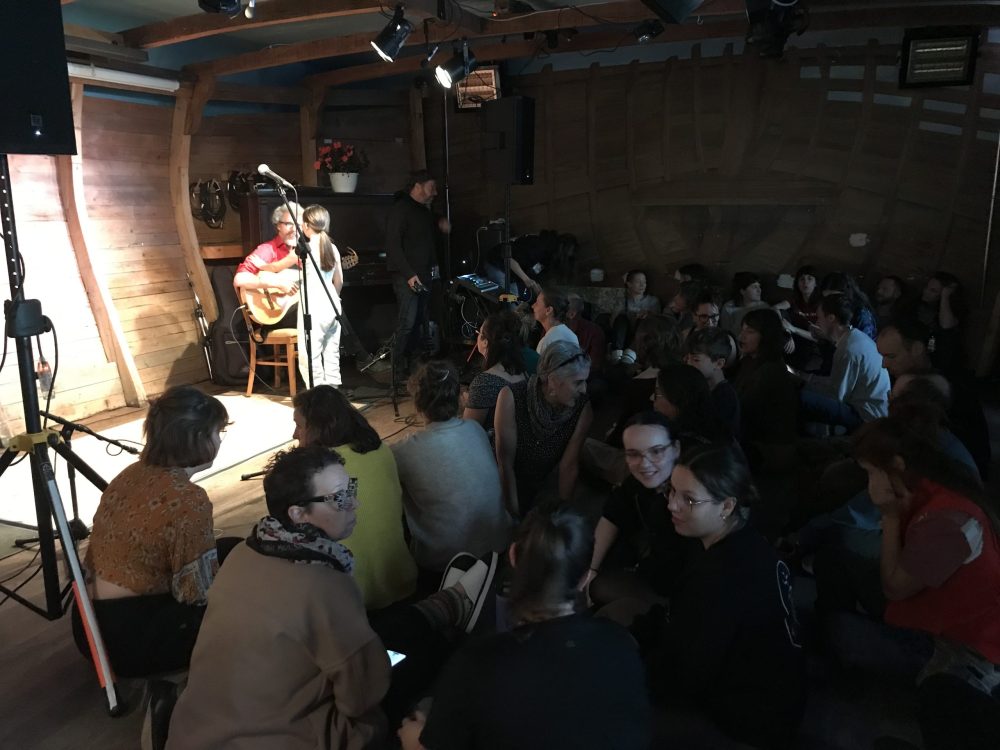 Une trentaine de personnes assises au sol devant une petit scène avec un micro
