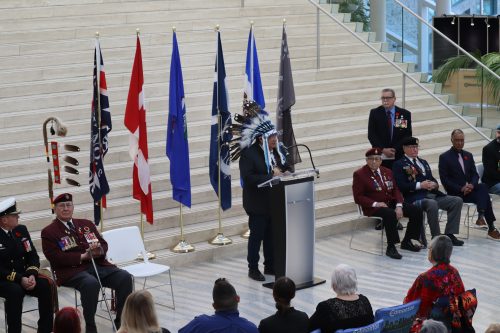 Chief Desmond Bull of the Louis Bull Tribe and the Treaty 6 Confederacy stands at the podium in Edmonton's City Hall, offering remarks on the city's proclamation. There are lots of people on his sides, and there are flags on the staircase behind him. Photo was taken inside.