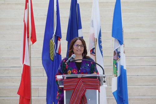 President Andrea Sandmaier of the Otipemisiwak Métis Government of the Métis Nation within Alberta delivers her remarks at Edmonton City Hall. The Canadian, Albertan, Metis, Metis Nation of Alberta, and Edmontonian flags stand behind her.