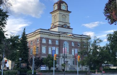 L'Hôtel de ville devant un ciel bleu avec le drapeau franco-manitobain dans le vent.