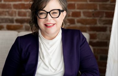 A portrait of Sheena Howard. She is sitting down and there is a red brick wall behind her. She wears a white shirt under a dark purple blazer and wears glasses.