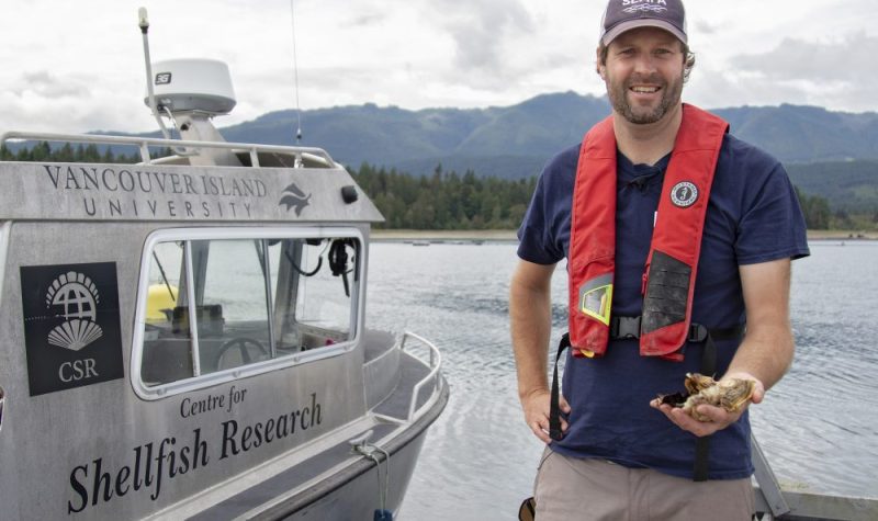 A man wearing a personal flotation device holds a shellfish in front of a boat reading Vancouver Island University Centre for Shellfish Research.