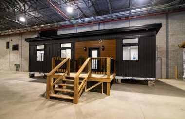 A small dark brown model home sits in a warehouse space with a wooden walkway in front of the home leading up to the front entrance.