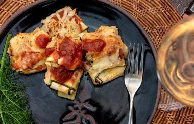 A dish with red tomato sauce topping sits on a black plate next to a glass of white wine.