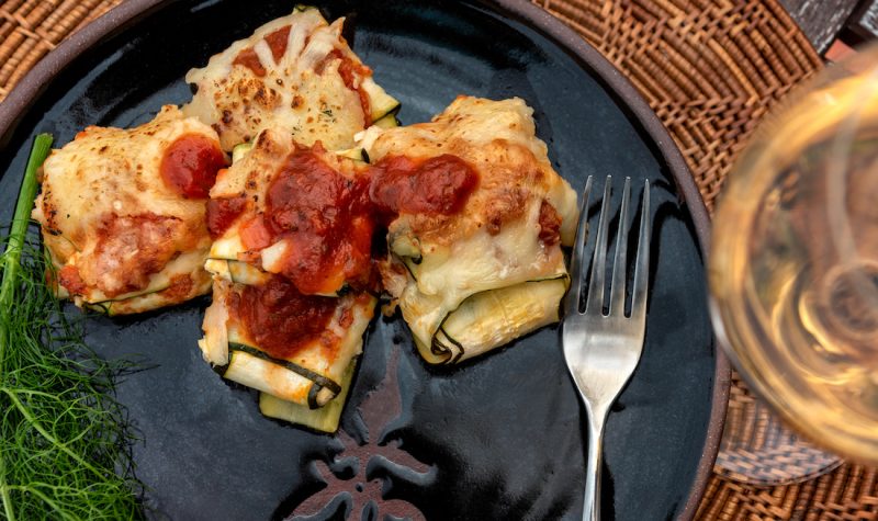 A dish with red tomato sauce topping sits on a black plate next to a glass of white wine.