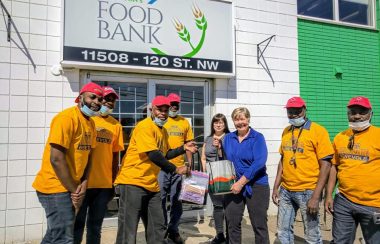 Six hommes habillés en jaune avec des casquettes rouges ainsi que deux femmes tenant des paniers en mains devant un bâtiment où c’est écrit Edmonton Food Bank