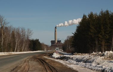 White smoke blows rightward from the smokestack of a power plant at the end of a road surrounded by trees; the land is partially covered by snow.