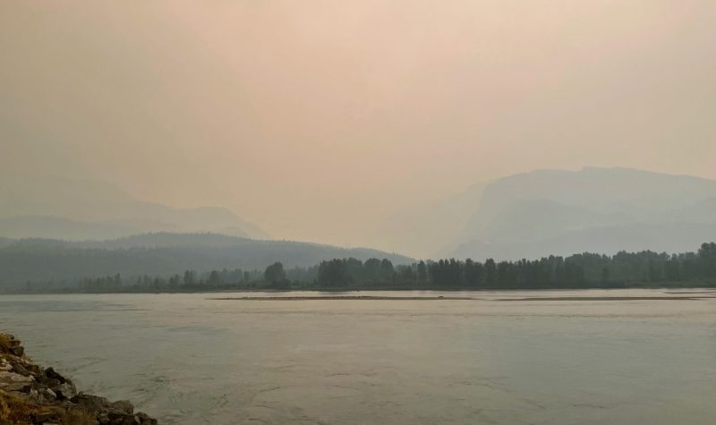 A wide angle photo of a river framed by riverbanks on either side. There are two mountains in the distance shrouded in grey smoke.