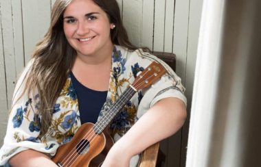 Jeune fille aux cheveux bruns assise sur une chaise avec une petite guitare entre les bras.