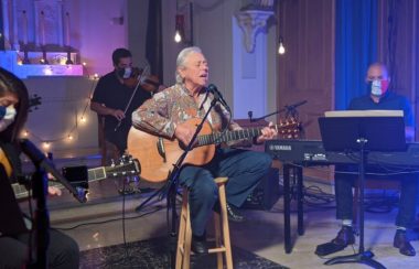 Une dame assise sur un banc jouant de la guitare et chantant avec des musiciens en arrière scène.