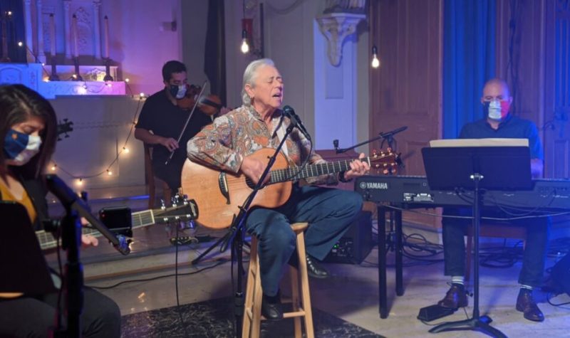 Une dame assise sur un banc jouant de la guitare et chantant avec des musiciens en arrière scène.