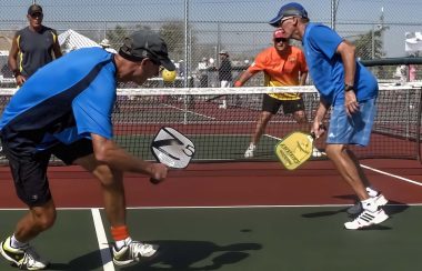 4 personnes jouent au Picleball, 2 personnes de chaque coté d'un filet, ils sont en plein action, ils ont des tenues de sport et des raquettes plus petite que des raquettes de tennis. Le temps est beau et ils sont concentrés.