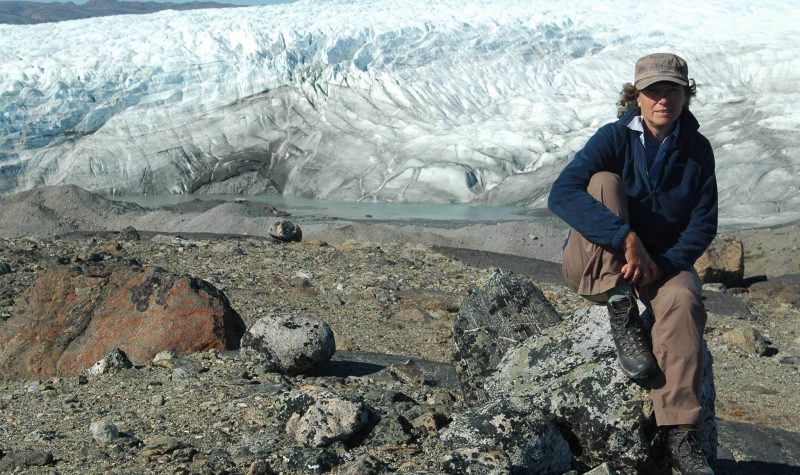 A women in hiking clothes sits in front of a large glacier.