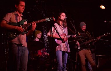 Jemma Hicken performs live with musicians on either side of her a lit stage.
