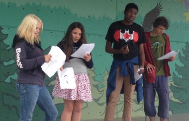 Four youth read from scripts upon a stage.