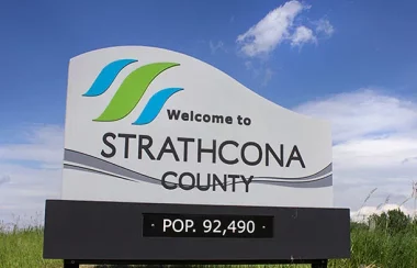 The welcome sign you see when coming into Strathcona County, with the logo on top, and the population on the bottom. Weather is partly cloudy.