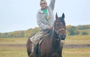An Indigenous man with mohawk and sunglasses raising fist towards the sky while sitting on a horse.
