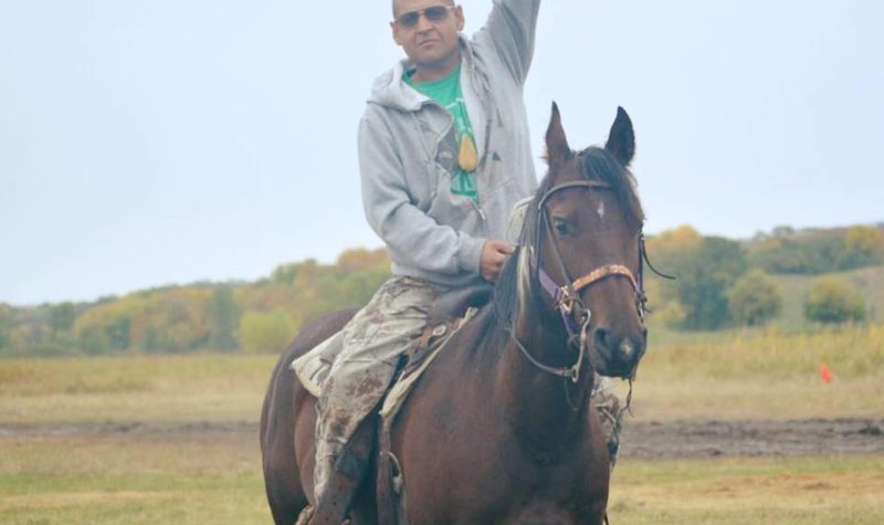 An Indigenous man with mohawk and sunglasses raising fist towards the sky while sitting on a horse.