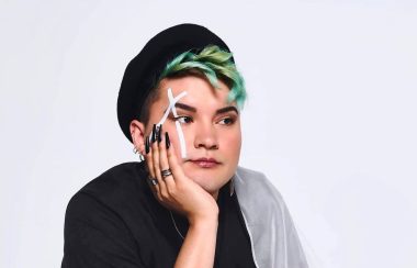 A studio portrait of Kairyn Potts, squatting down with an opaque flag draped down his left shoulder. He also has a white X painted across his right eye, while wearing a hat a little far back on his head showing his green hair.