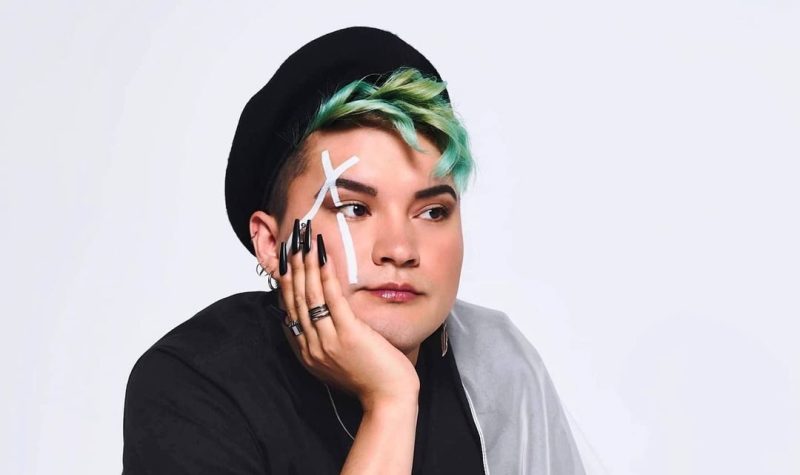 A studio portrait of Kairyn Potts, squatting down with an opaque flag draped down his left shoulder. He also has a white X painted across his right eye, while wearing a hat a little far back on his head showing his green hair.