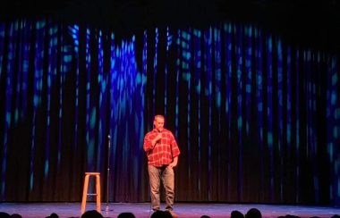 Humoriste avec chemise orange et culottes brunes en spectacle à côté d,un banc et devant une foule.