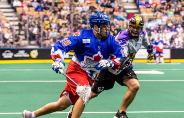 A closeup of two lacrosse players, on the playing field in front of an audience. One is wearing a red white and blue uniform, the other is wearing purple and black.