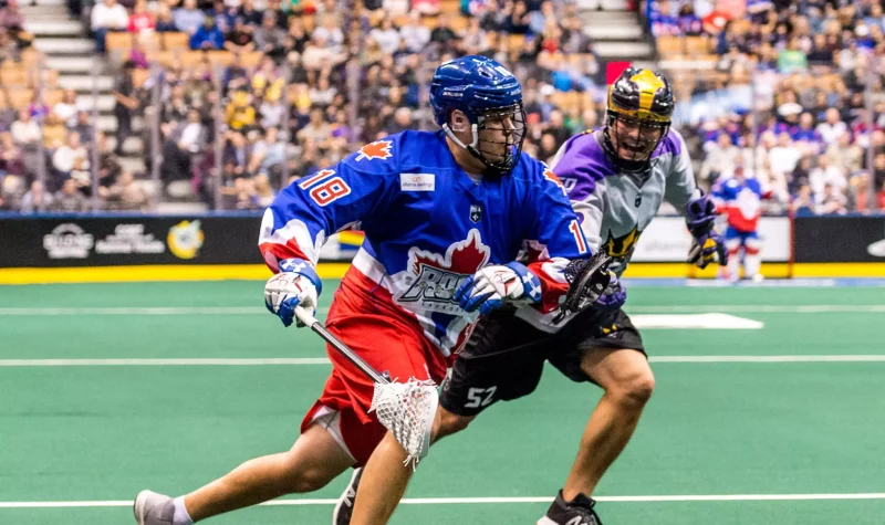 A closeup of two lacrosse players, on the playing field in front of an audience. One is wearing a red white and blue uniform, the other is wearing purple and black.