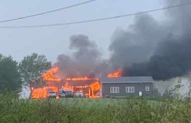 A house is seen across a field. The house is on fire with four vehicles parked in front. It stands next to a grey garage and grey smoke billows from the house and to the right