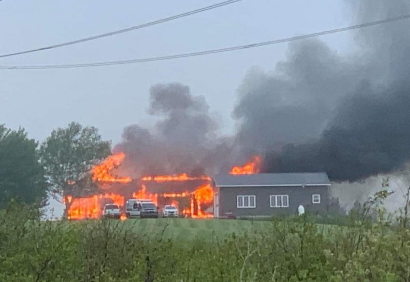 A house is seen across a field. The house is on fire with four vehicles parked in front. It stands next to a grey garage and grey smoke billows from the house and to the right