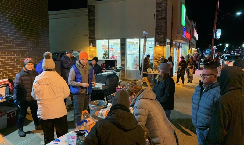 A crowd of people gathered outside, wearing hats and coats, around tables with someone working a bbq.