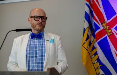 A man wearing a blue checkered shirt, a blue bow-tie and a white jacket stands at a lecture with the BC flag in the background.