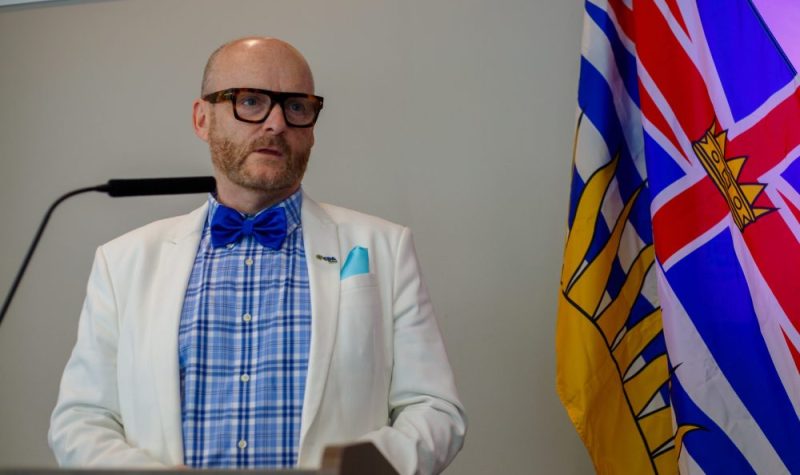 A man wearing a blue checkered shirt, a blue bow-tie and a white jacket stands at a lecture with the BC flag in the background.