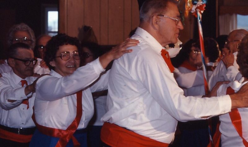 Hommes et femmes habilés en blanc, rouge, bleu et noir dansant dans un cercle avec les mains sur l'épaule.